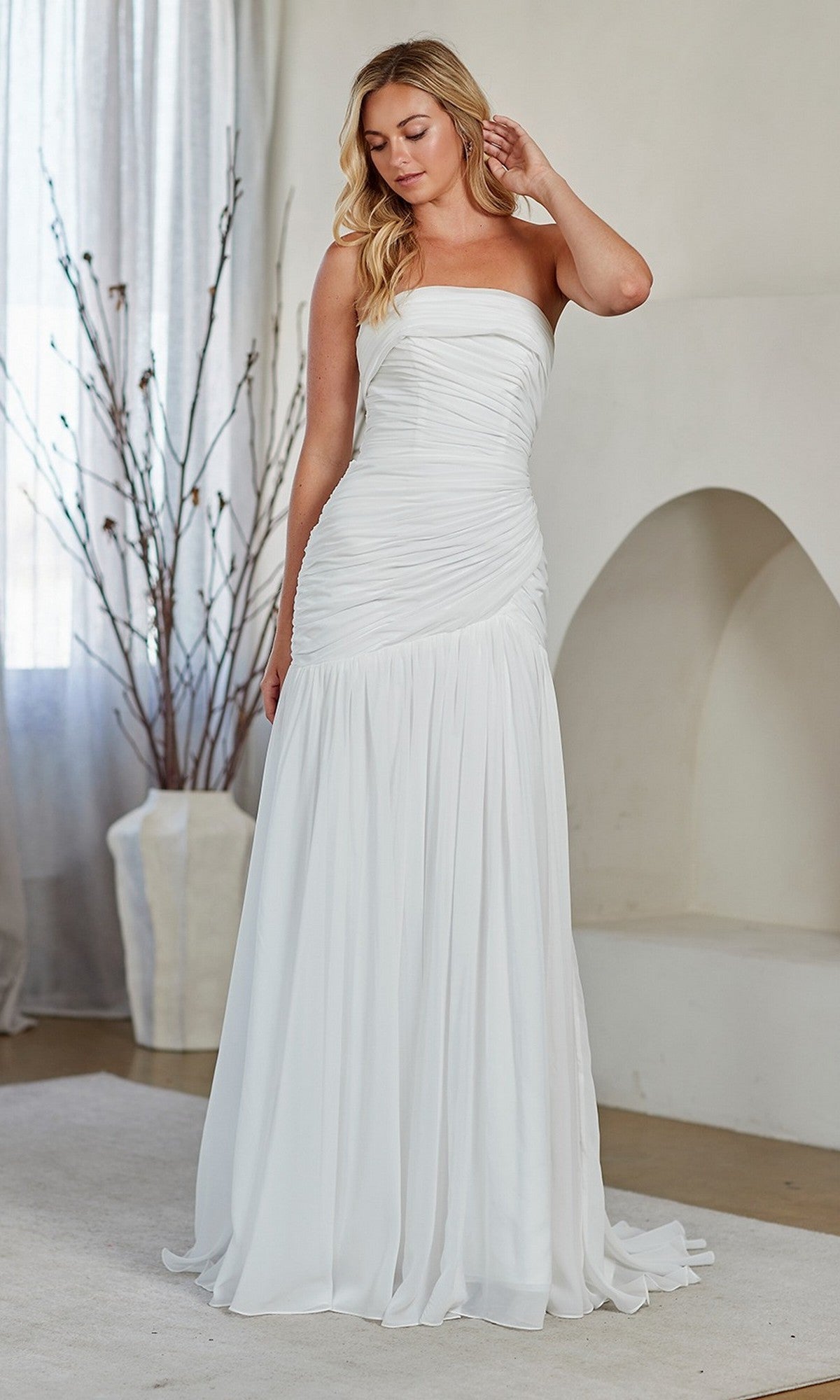 A woman stands indoors on a light rug, wearing the Amelia Couture 6132 long prom dress with a strapless neckline and flattering ruching, framed by a large vase of branches and a white arched alcove in the background.